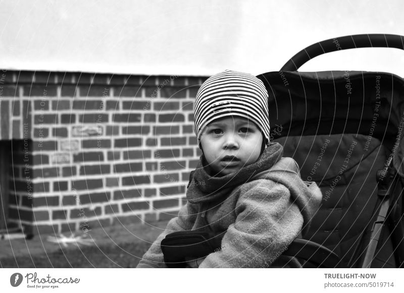 Energiekrise? Ein kaum zwei Jahre altes Kind sitzt warm eingepackt mit Mütze, Schal, Fleece-Jacke im seinem Buggy und blickt sehr ernst und fragend direkt in die Kamera