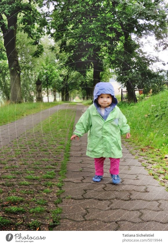 Kleines Mädchen läuft Allee entlang regnerisch Regen Regenjacke Kind Gummistiefel Kaputze grün Mensch Außenaufnahme schlechtes Wetter nass Kindheit spielen