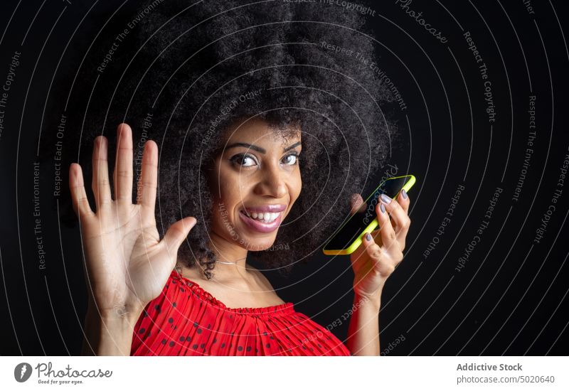 Lächelnde kubanische Frau mit Smartphone grüßt auf schwarzem Hintergrund Hallo Gruß feminin Hand erhoben freundlich Afro-Look Porträt Funktelefon Apparatur