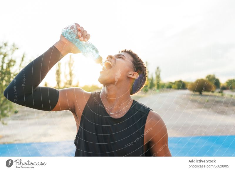 Schwarzer Sportler trinkt auf einem Spielplatz Wasser aus einer Flasche durstig Gesundheit trinken Training jung männlich Athlet Sportbekleidung Mann aktiv