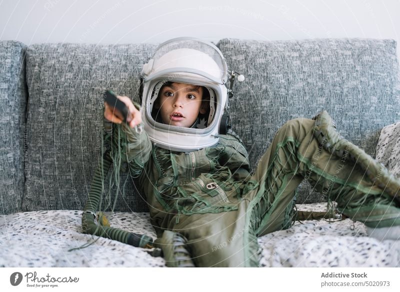 Erstauntes Kind, das mit einer echten Astronautenuniform auf der Couch sitzt und eine TV-Fernbedienung benutzt Hintergrund Junge Kindheit Konzept Tracht