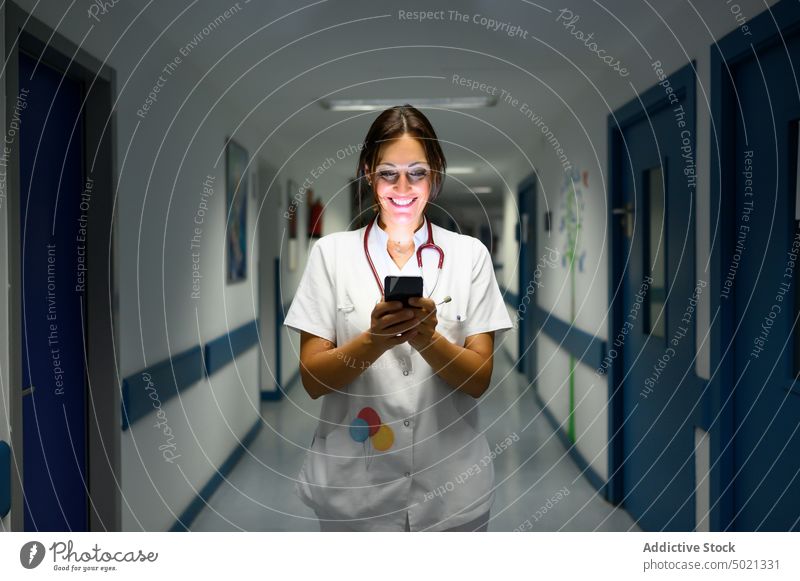 Fröhliche Kinderärztin mit Smartphone auf dem Krankenhausflur benutzend Gang Spaziergang Lächeln Uniform Frau Pause Arzt Arbeit Job Gerät Apparatur dunkel