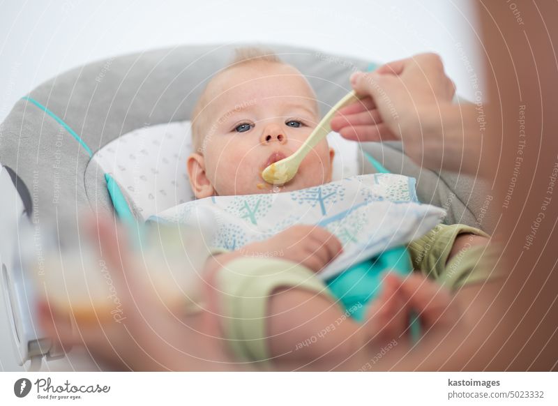 Mutter Löffel füttern ihr Baby Junge Kleinkind Kind in Babystuhl mit Fruchtpüree. Baby feste Nahrung Einführung Konzept. Gesundheit Ernährung Essen Lebensmittel
