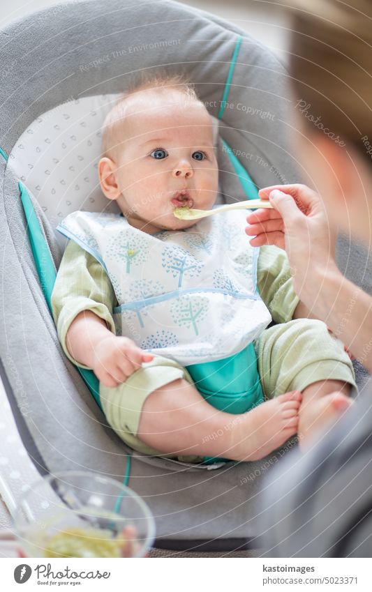 Mutter Löffel füttern ihr Baby Junge Kleinkind Kind in Babystuhl mit Fruchtpüree. Baby feste Nahrung Einführung Konzept. Gesundheit Ernährung Essen Lebensmittel