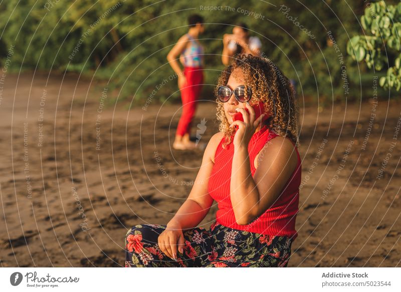 Ethnische Frau, die am Strand mit ihrem Smartphone spricht Sommer reden Stil Sand farbenfroh Kommunizieren Telefon ethnisch jung krause Haare Sonnenbrille