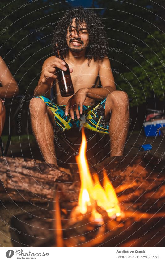 Junger ethnischer Mann trinkt Bier mit Freunden am Lagerfeuer Freudenfeuer Strand trinken heiter Flasche Alkohol Glück Abend Picknick jung ohne Hemd