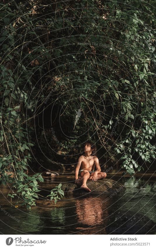 Verträumter Junge, der auf einem Stein am Flussufer im Wald ruht verträumt Rüssel sich[Akk] entspannen positiv Kind Küste üppig (Wuchs) Waldgebiet Natur Ufer