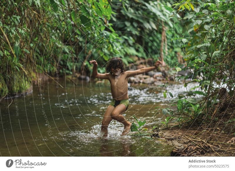 Glücklicher ethnischer Junge, der in einen Bach im Wald springt springen Fluss freudig Spaß haben Kind sorgenfrei heiter Aktivität üppig (Wuchs) seicht