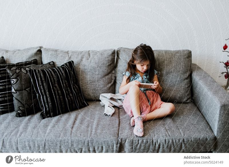 Niedliches ethnisches Kind mit Smartphone auf gemütlicher Couch zu Hause benutzend spielen sich[Akk] entspannen Sofa Komfort online Internet Videospiel