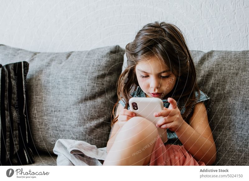 Niedliches ethnisches Kind mit Smartphone auf gemütlicher Couch zu Hause benutzend spielen sich[Akk] entspannen Sofa Komfort online Internet Videospiel