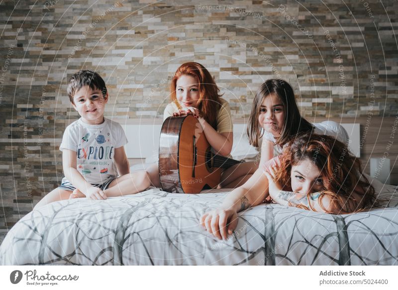 Lächelnde Frauen mit Gitarre auf dem Bett sitzend mit Kindern Spaß Freundschaft im Innenbereich alternativ Schlafzimmer heiter Kindheit Komfort gemütlich