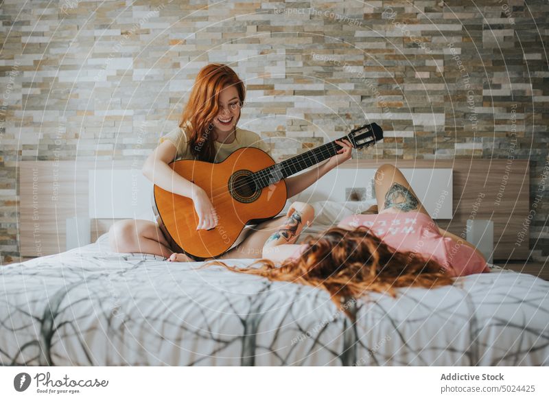 Lächelnde Frauen, die sich auf dem Bett ausruhen und auf der Gitarre spielen Musik Schlafzimmer im Innenbereich Freizeit akustisch alternativ Kunst heiter