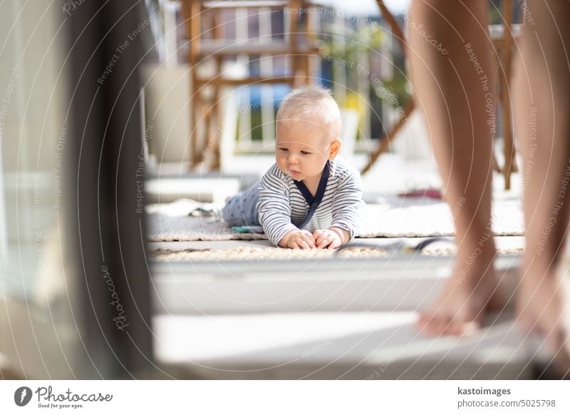 Niedliches kleines Baby, das mit Spielzeug im Freien auf der Terrasse im Sommer spielt und von seiner Mutter im Hintergrund beaufsichtigt wird. Säugling