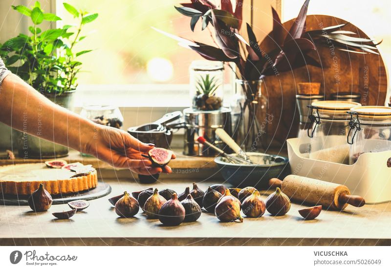 Frauenhand, die Feigen auf dem Küchentisch mit Backwerkzeugen im Fensterhintergrund hält. Lebensstil Hand Beteiligung Tisch Lifestyle im Innenbereich