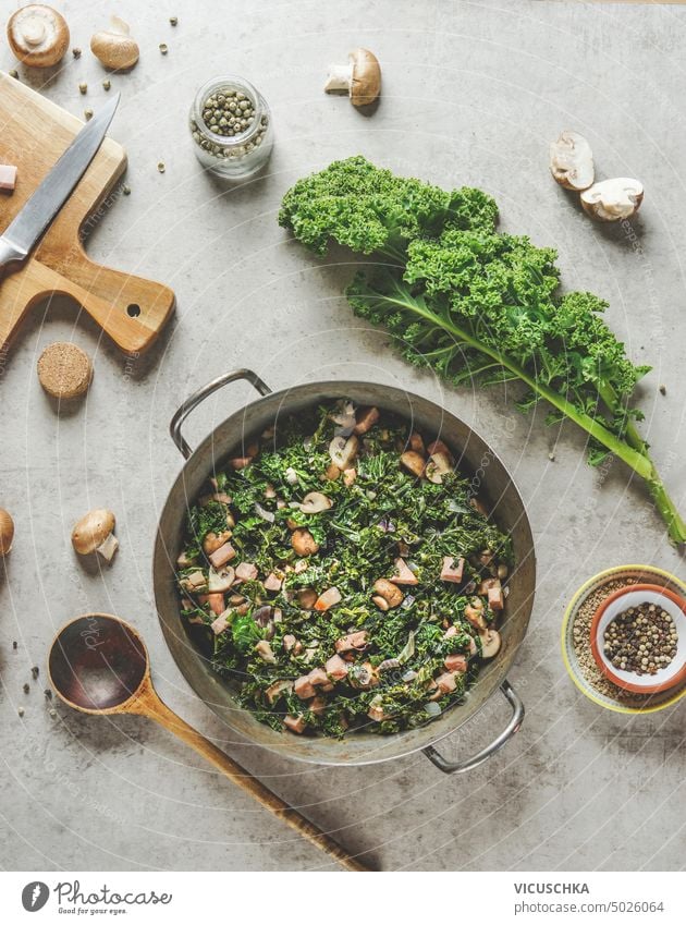 Kochtopf mit gedünstetem Grünkohl mit Pilzen auf Küchentisch mit Löffel und Zutaten, Ansicht von oben Kale Tisch Draufsicht Essen zubereiten lecker Gemüse