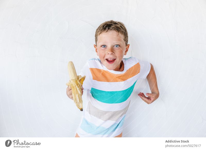 Fröhliches süßes Kind isst Banane und lächelnd lehnt an der weißen Wand Lächeln positiv Frucht Junge Glück Vitamin bezaubernd Porträt frisch Kindheit froh