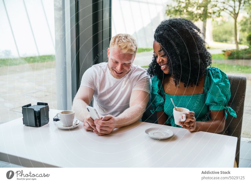 Fröhliches gemischtrassiges Paar sitzt in einem Café und benutzt ein Mobiltelefon Termin & Datum Kantine Partnerschaft Sitzung Browsen Zusammensein Freund