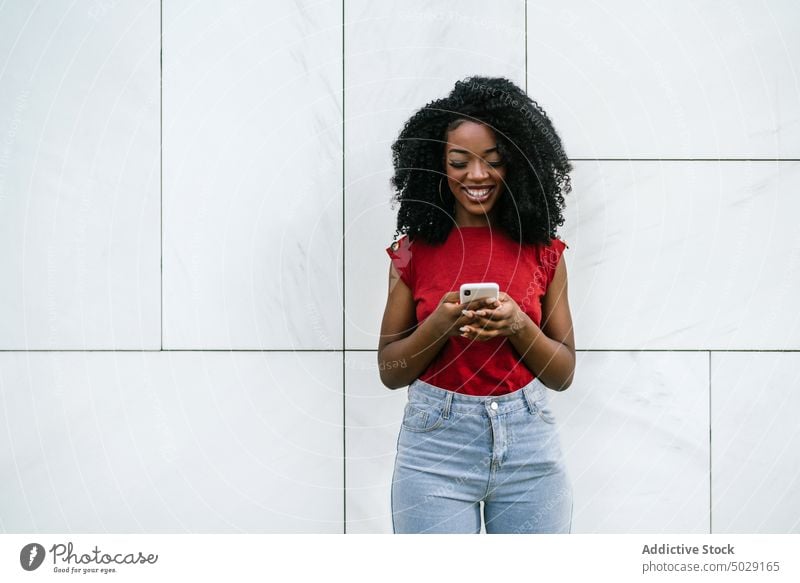 Fröhliche afroamerikanische Frau, die in der Nähe der Wand auf ihrem Smartphone surft heiter Lächeln Browsen Funktelefon Telefonanruf Straße Glück Stil Gerät