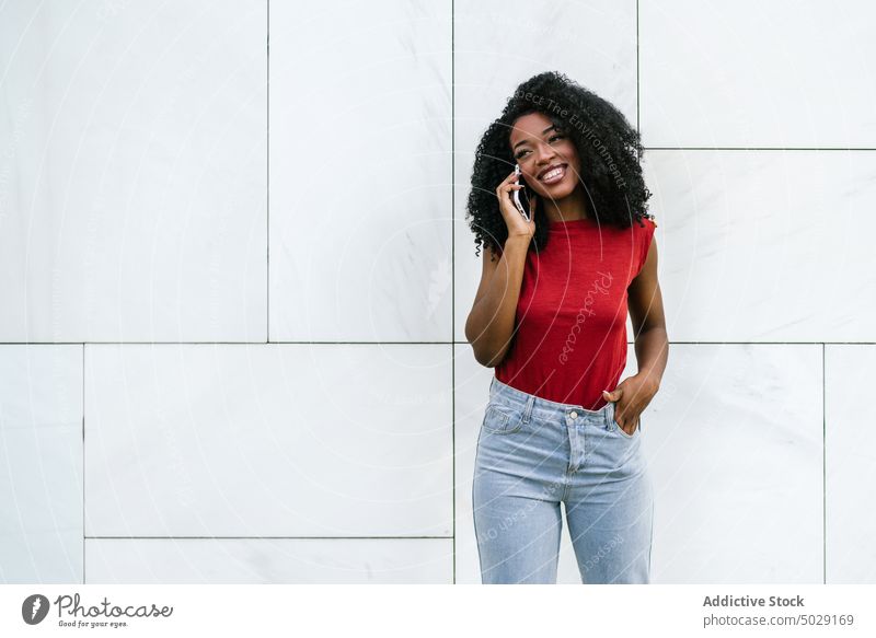Fröhliche afroamerikanische Frau, die in der Nähe der Wand mit einem Smartphone spricht heiter Lächeln Funktelefon reden Hand-in-Tasche sprechen Telefonanruf
