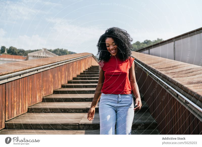 Fröhliche ethnische Frau, die auf einer Holztreppe geht Lächeln Glück hölzern Treppenhaus Spaziergang Großstadt Straße Outfit Afro-Look urban Afroamerikaner