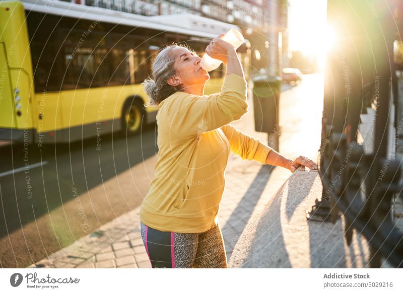 Ältere Sportlerin macht Pause auf der Straße trinken Fitness Training Sonnenaufgang Flasche Wasser Zaun Frau älter Senior gealtert Wellness Energie Kraft