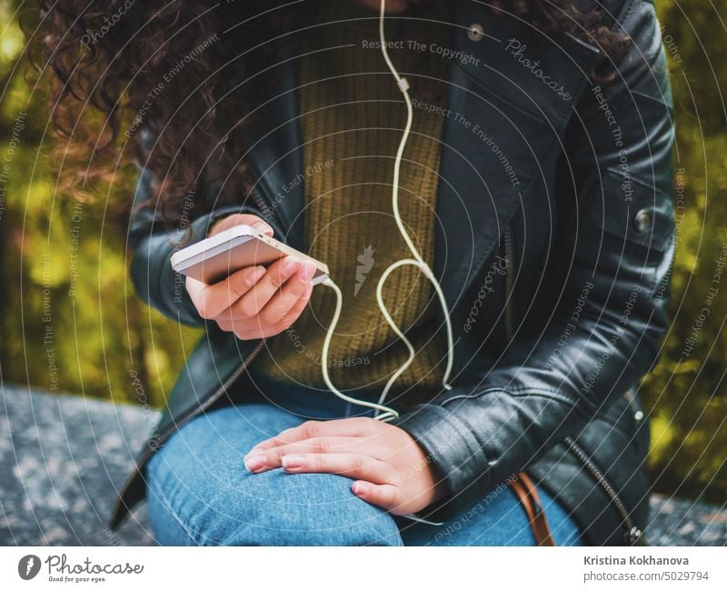 Hübsches junges lateinisches Mädchen mit schwarzem lockigem langem Haar hört Musik mit Kopfhörern und Smartphone oder Player auf einer verlassenen Herbststraße. Frau genießt den Moment.
