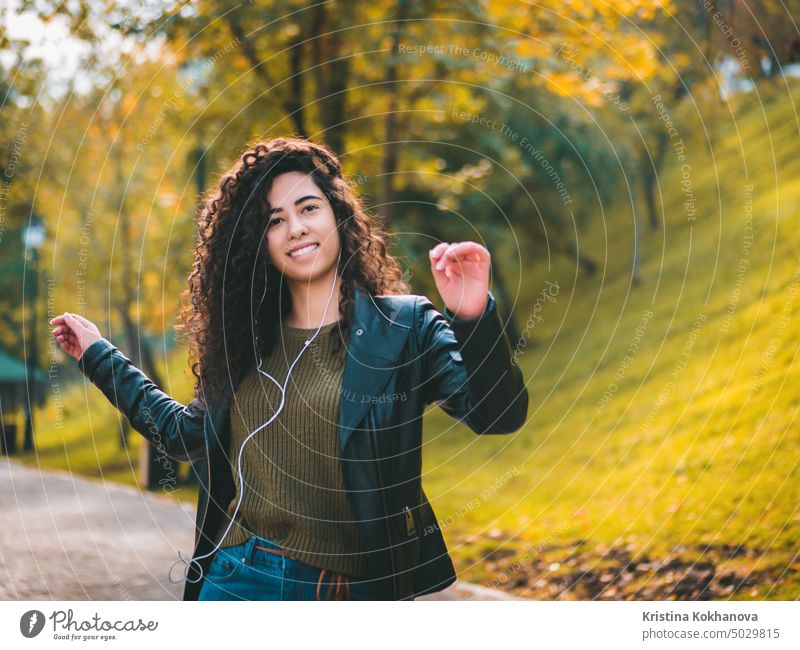 Glückliche afrikanische junge Frau mit lockigem Haar hört Musik über Kopfhörer. Hispanic Hipster Mädchen tanzen zu Rhythmus und singen entlang Melodie im Herbst Park.