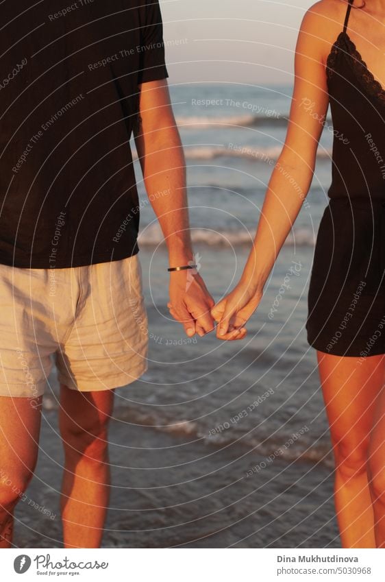 Unbekanntes Paar hält sich am Strand an den Händen. Mann und Frau zusammen mit Wellen des Meeres auf dem Hintergrund. Nahaufnahme. Gebräunte Haut bei Sonnenuntergang. Liebe und Beziehungen. Sommer Liebesgeschichte von jungen Erwachsenen. Millennial im Urlaub.