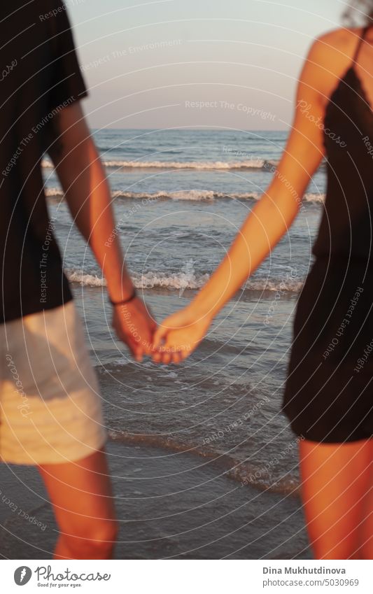 Unbekanntes Paar hält sich am Strand an den Händen. Mann und Frau zusammen mit Wellen des Meeres auf dem Hintergrund. Nahaufnahme. Gebräunte Haut bei Sonnenuntergang. Liebe und Beziehungen. Sommer Liebesgeschichte von jungen Erwachsenen. Millennial im Urlaub.