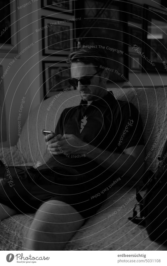 Smarter Typ sitzt breitbeinig im Dunkeln mit Sonnenbrille auf Sofa und blickt gebannt auf sein Smartphone [vertieft oder app-hängig?] Mann lässig cool abhängig