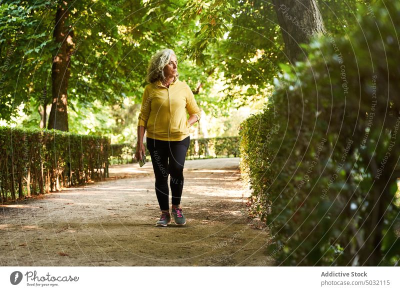 Ältere Sportlerin beim Spaziergang im Park Weg Training Herz Fitness Sommer Aktivität Gasse Frau Senior gealtert älter Buchse Strauch üppig (Wuchs) Athlet