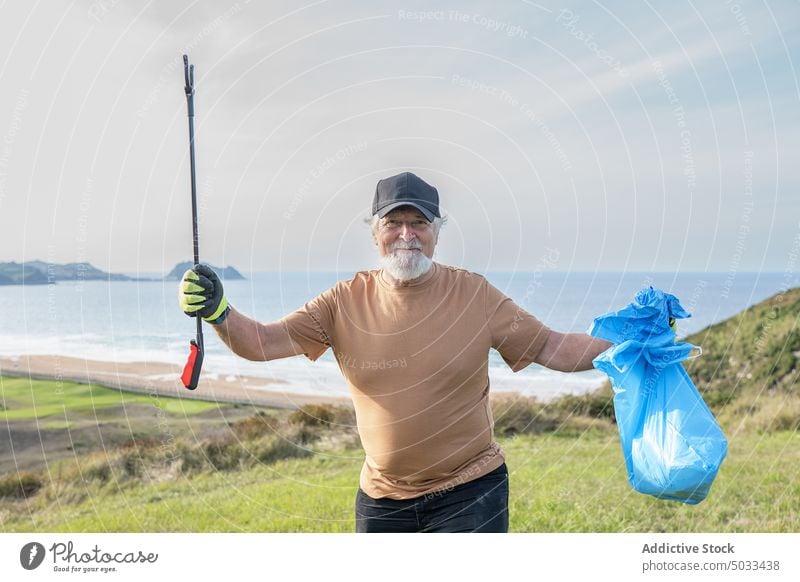 Positiver älterer Mann mit Müllzange und Sack auf einem Hügel stehend Rentnerin Lächeln Freiwilliger Tasche Umwelt Natur abholen MEER verschmutzen Ökologie