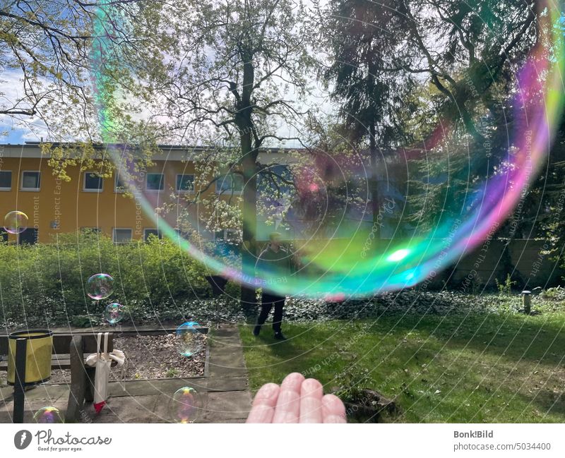 Ein Mensch lässt Seifenblasen fliegen. Im Vordergrund eine Hand und eine riesige, in Regenbogenfarben schillernde Seidenblase, die langsam davon schwebt. Im Hintergrund weitere, die schon eine Strecke geflogen sind. Garten, Spielplatz.