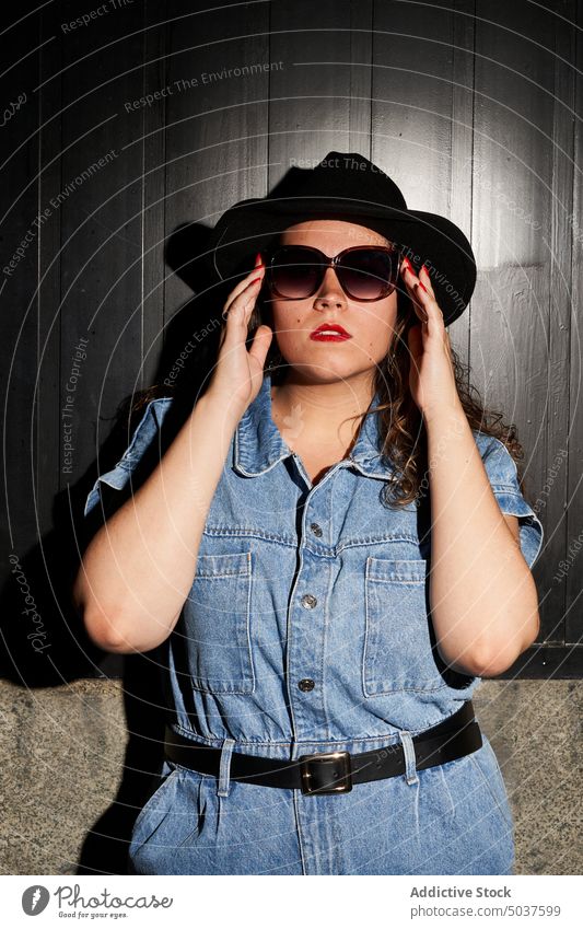 Stilvolle junge Frau, die eine Sonnenbrille berührt ausrichten Vorschein Wand Hut lässig selbstbewusst Porträt Madrid Spanien Brille Persönlichkeit Jeansstoff