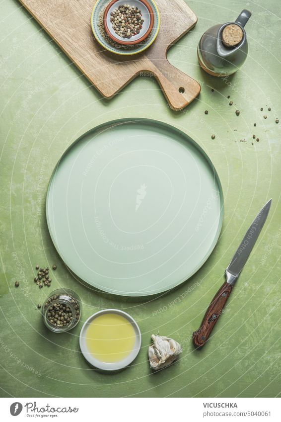 Grüner Lebensmittelhintergrund mit leerem Teller, Schneidebrett, Messer, Speiseöl und Gewürzen, Ansicht von oben. Rahmen grün Hintergrund Öl Draufsicht