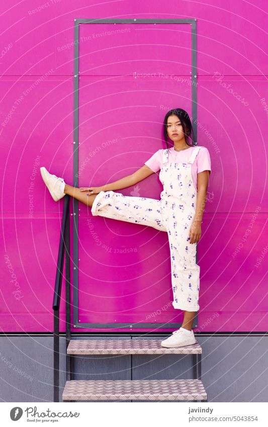 Asiatische Frau, die mit ernster Miene in die Kamera blickt und ein Bein über das Geländer in der Nähe der rosa Wand hebt. asiatisch Chinesisch modisch urban