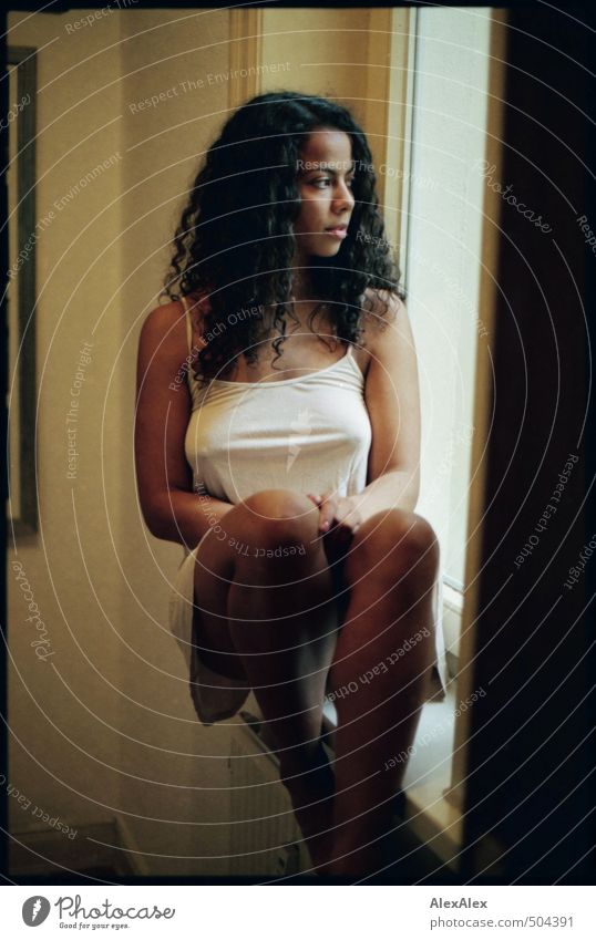 Eine junge, schöne Frau sitzt innen in einem Fensterrahmen - analoge Fotografie junge Frau langhaarig lockig Locken sportlich schlank Beine barfuß Raum Innen