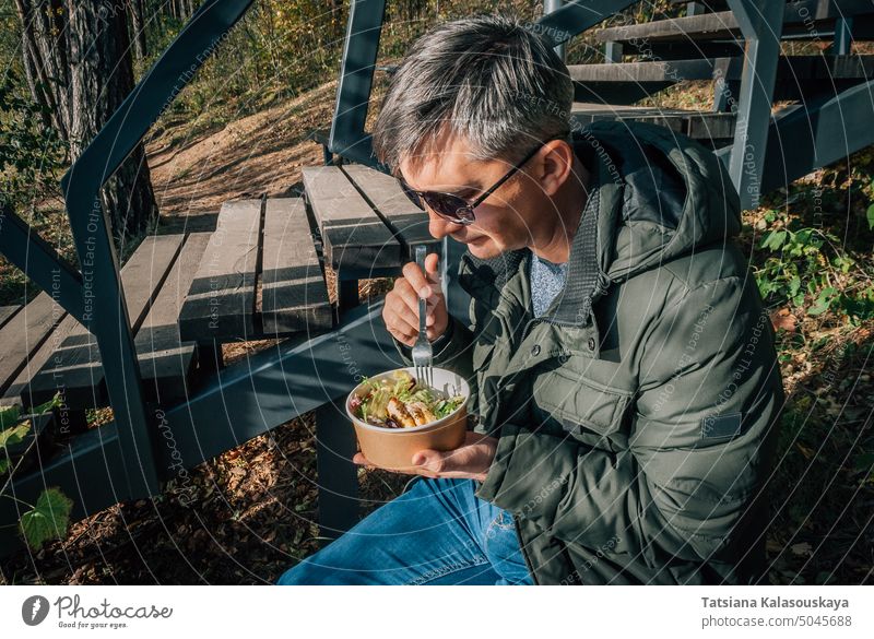 Ein Mann isst bei einem Herbstspaziergang in der Natur einen Salat aus einer Einweg-Papierschüssel im Freien Park Lifestyle Lebensmittel Mittagessen wegnehmen