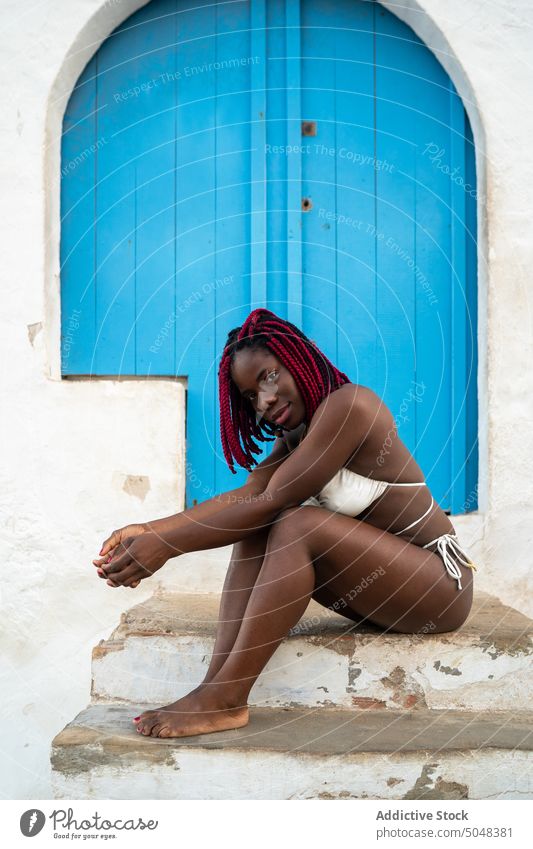 Stilvolle Frau sitzt vor einem Haus mit blauer Tür Schritt Tourist Bikini Sommer Urlaub sich[Akk] entspannen sinnlich Rastalocken Outfit selbstbewusst trendy