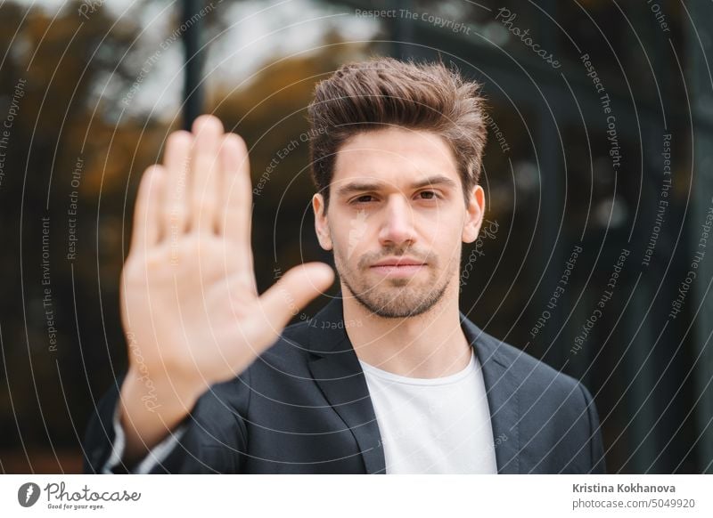 Porträt eines jungen Geschäftsmannes, der mit der Hand eine ablehnende Geste macht: Verweigerungszeichen, kein Zeichen, negative Geste, schließt die Kamera mit der Hand, professioneller männlicher Manager mit Anzugjacke.