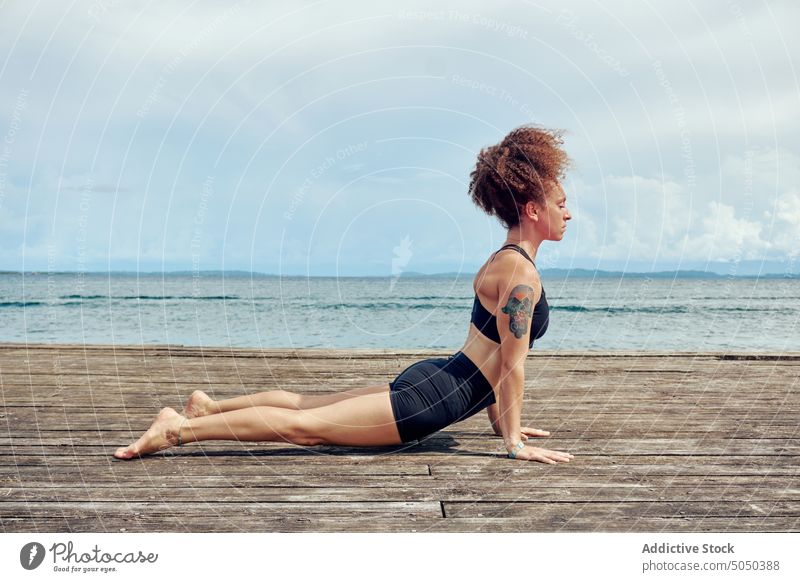 Ruhige ethnische Frau in Yogastellung auf dem Pier üben Kai MEER Sommer Gesundheit bhujangasana Kobra-Pose Natur Wellness Harmonie Atem Ufer Hafengebiet Energie