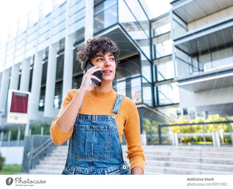 Zufriedene Frau bei einem Telefongespräch vor einem Gebäude an einem sonnigen Tag Smartphone benutzend Audio reden sprechen Anruf Straße Sommer urban Stil