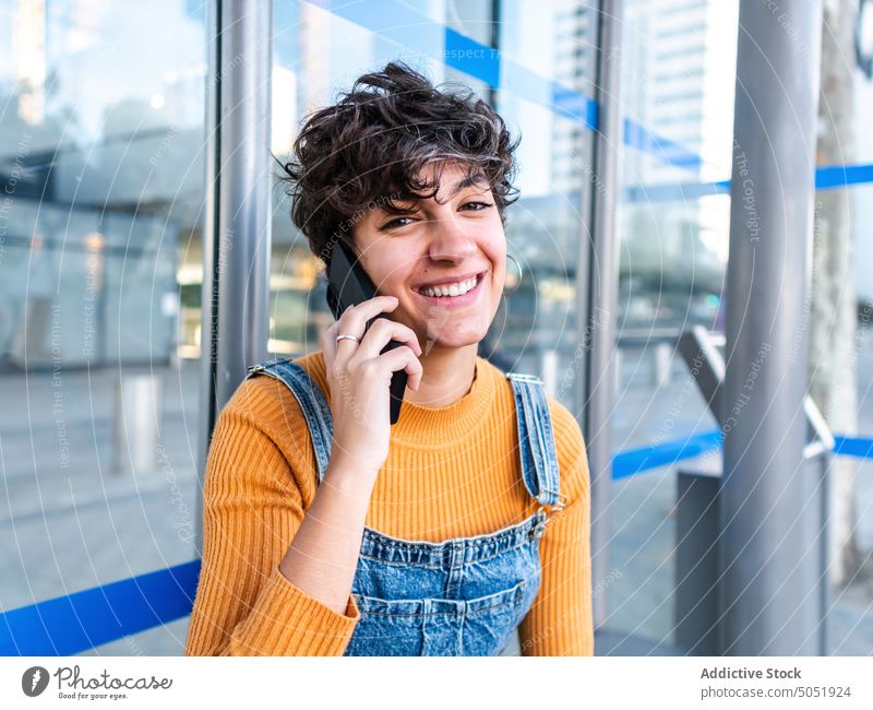 Fröhliche Frau spricht am Telefon in der Nähe eines modernen Glasgebäudes heiter Lächeln reden Smartphone Telefonanruf Straße urban Gebäude Glaswand Mobile