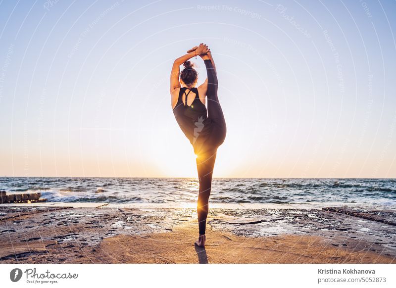 Schlanke Frau in schwarzem Bodysuit übt Yoga in der Nähe von Meer oder Ozean bei Sonnenaufgang Licht. Flexibilität, Stretching, Fitness, gesunder Lebensstil.