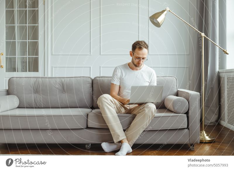 Junger europäischer Mann Freiberufler sitzt auf der bequemen Couch im Wohnzimmer, arbeitet am Laptop Sofa heimwärts Arbeit Technik & Technologie Computer lässig