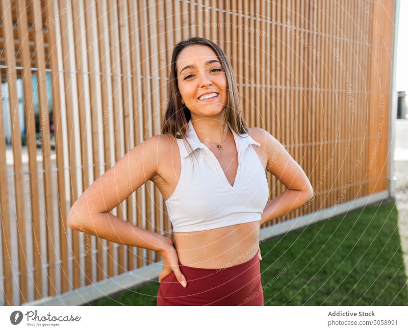 Gut gelaunte Sportlerin in der Nähe eines Holzzauns Lächeln Hand auf der Taille Zaun hölzern Sportbekleidung Athlet positiv Park Gras Gesundheit Pause Frau jung