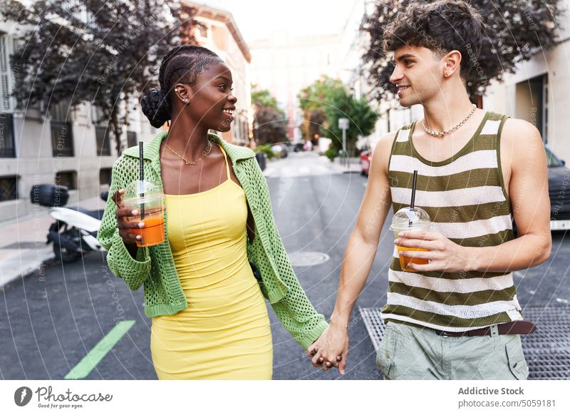 Verliebtes junges multiethnisches Paar hält sich an den Händen und trinkt Säfte beim Gehen auf der Straße Händchenhalten Lächeln Spaziergang Saft Partnerschaft