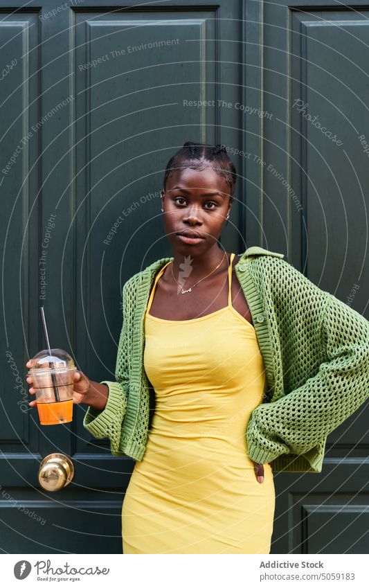 Stilvolle junge schwarze Frau mit einer Tasse Saft zum Mitnehmen, die in der Nähe der Tür steht selbstbewusst Straße Imbissbude Hand auf der Taille ernst