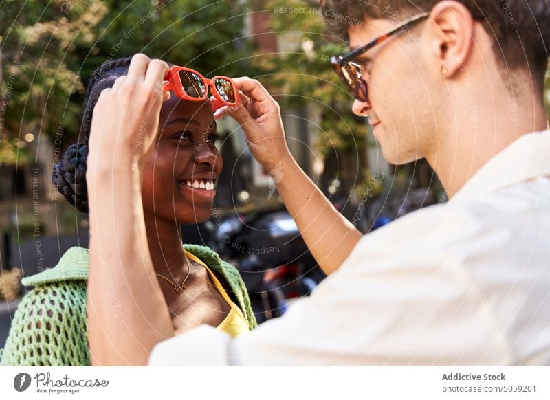 Liebevoller ethnischer Mann passt die Sonnenbrille seiner lächelnden Freundin auf der Straße an Paar Lächeln Partnerschaft ausrichten positiv Stil romantisch