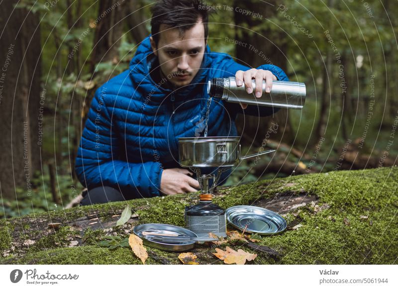 Ein Mann in einer blauen Daunenjacke kocht mitten in der Wildnis auf einem tragbaren Kocher und in einem ultraleichten Topf aus Edelstahl Wasser für Tee und Essen. Lebensstil beim Wandern